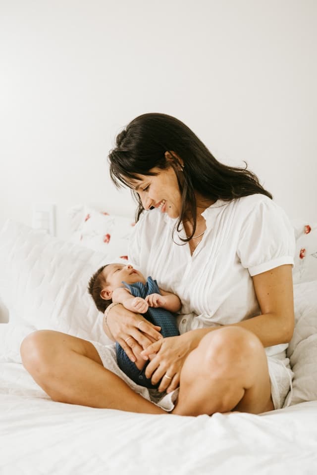 A woman wearing a white dress holds a newborn on a bed.