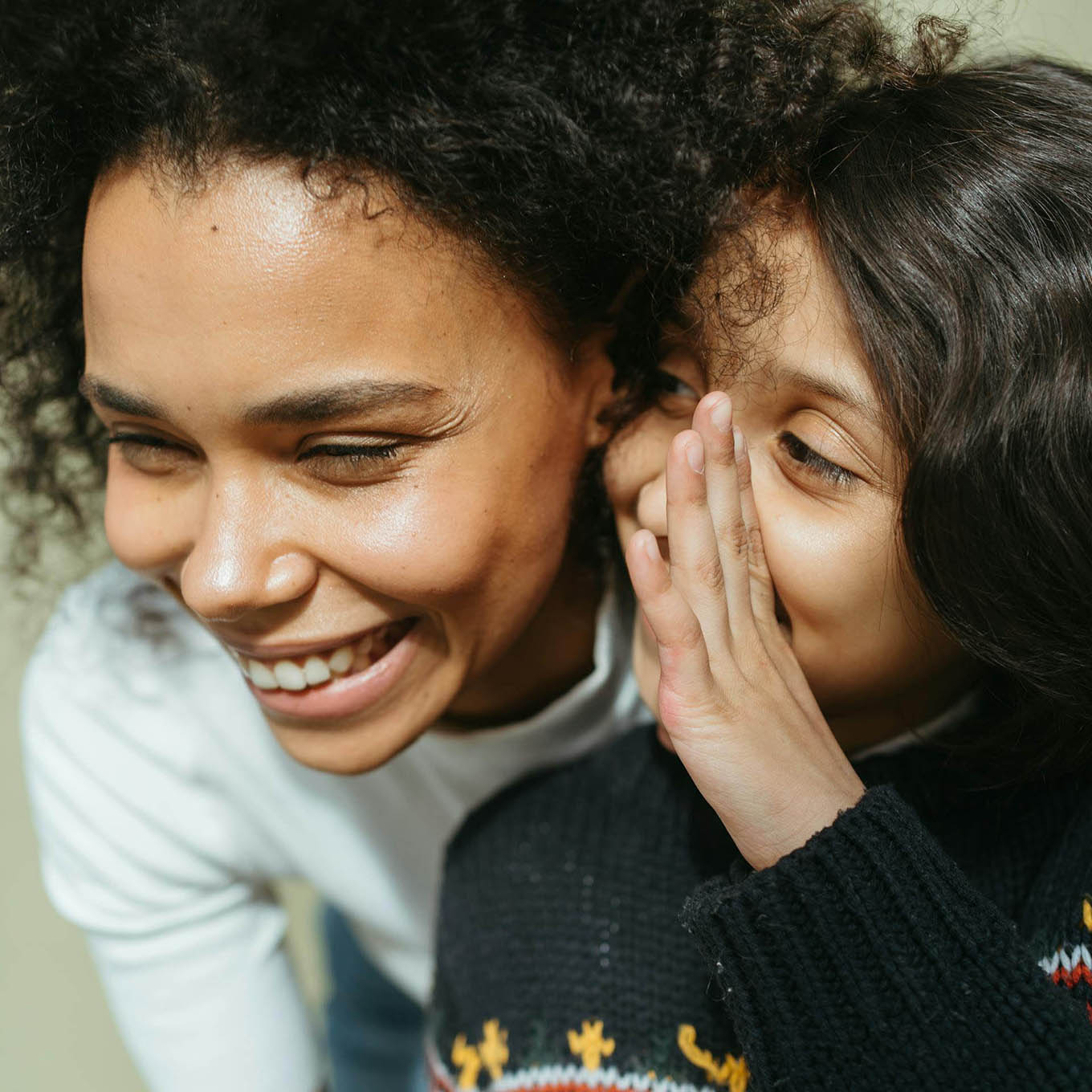 A curly-haired woman laughs while a child whispers in her ear.