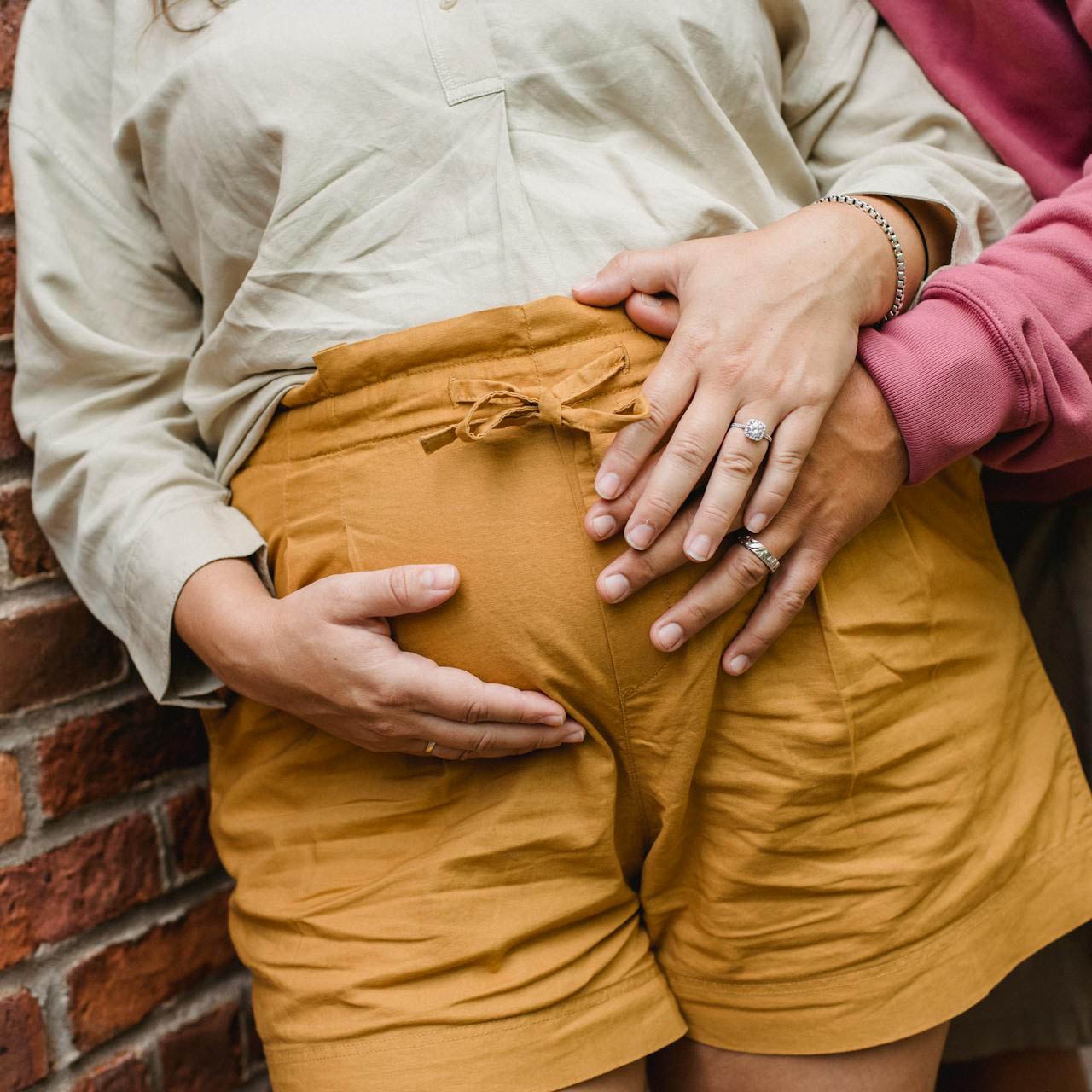 A couple stands against a brick wall while holding the woman's pregnancy bump.