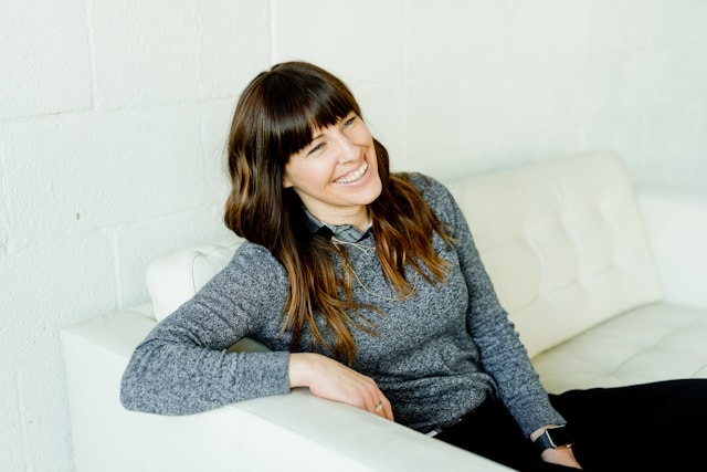 A woman with brown hair sits on a white couch and smiles.