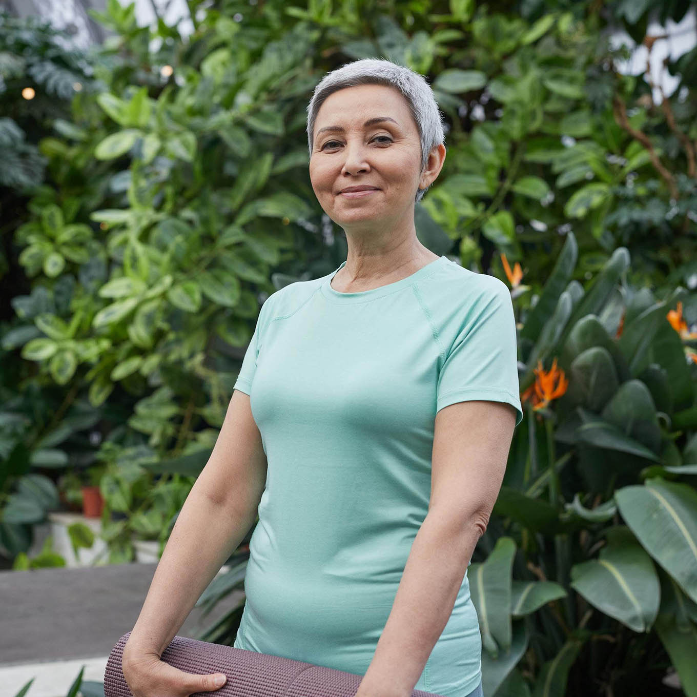 A middle-aged woman smiles and holds a yoga mat while standing in a lush garden.